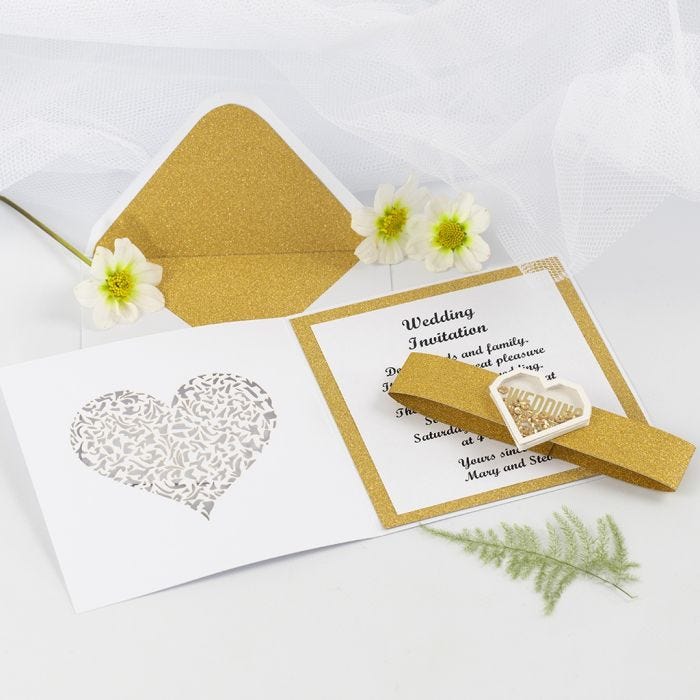 Una invitación de boda con papel de diseño dorado y una pegatina shaker en forma de corazón.