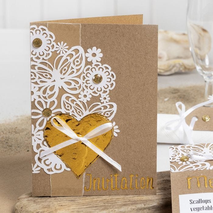 Una invitación decorada con encaje de cartulina, corazón de Deco Foil y pedrería.